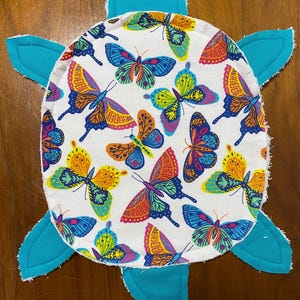 May include: A handmade turtle-shaped item with a white center patterned with colorful butterflies. The turtle's head and flipper-shaped appendages are a solid turquoise blue. The butterfly wings feature shades of orange, yellow, blue, and purple.