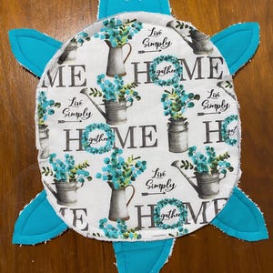 May include: A turtle-shaped decorative item with a white center featuring the word "HOME" and floral arrangements. The text "Live Simply" and "gather" are also present. The turtle's head and flippers are bright turquoise.