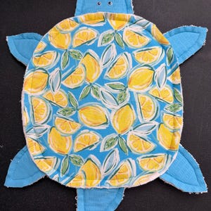 May include: A turtle-shaped fabric mat featuring a bright blue background with a repeating pattern of yellow lemon slices and green leaves. The mat has a quilted texture, a solid blue head with two small googly eyes, and four blue flippers.