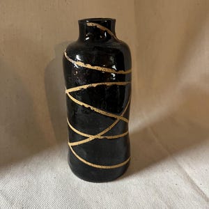 Può includere: Un vaso di ceramica nero con finitura lucida. Il vaso è decorato con linee dorate che si incrociano. Il vaso ha una forma cilindrica con una base leggermente più larga e un collo stretto. Il design è semplice ma elegante.