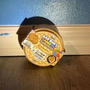 May include: A round, yellow Bao Bun's Rainbow Surprise toy container with a textured lid resembling a bamboo steamer. The label features the text Bao Bun's Rainbow Surprise, The Real Surprise Bao Bun, and colorful graphics of small food items.