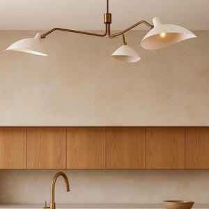 May include: A ceiling-mounted chandelier featuring a central brass rod and three adjustable arms extending outwards. Each arm holds a white, wide-brimmed, bowl-shaped shade with a warm light bulb visible inside, suspended above a kitchen counter.