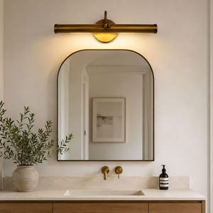 Pode incluir: Um conjunto de casa de banho com um espelho arqueado com uma moldura escura fina, uma luminária de parede horizontal em latão, uma bancada de pedra com lavatório embutido, torneiras de parede em latão e um vaso de cerâmica com folhagem.