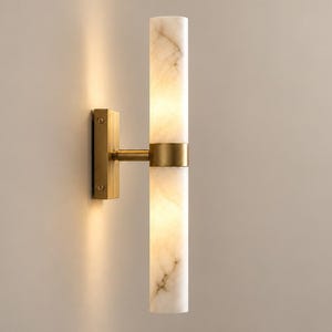 May include: A wall-mounted light fixture featuring two vertical cylindrical white marble shades with grey veining, separated and held by a central brushed brass band and connected to a rectangular brass wall plate by a horizontal arm.
