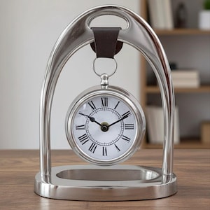 May include: A silver desk clock with a round face and Roman numerals. The clock is suspended within a curved, arch-shaped frame and has a brown leather strap at the top. The base is also silver. The clock is a decorative item.