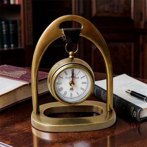 Op de afbeelding: Een messingkleurige bureauklok met een ronde wijzerplaat en Romeinse cijfers. De klok hangt aan een gebogen messing frame met een leren band. De wijzerplaat leest "Franklin Murphy Oxford". De klok staat op een houten bureau.