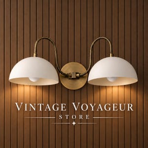 May include: A double wall sconce featuring two matte white dome shades attached to a central circular brass mounting plate by curved brass arms. The text VINTAGE VOYAGEUR STORE is centered below the light fixture against a vertical wood panel wall.