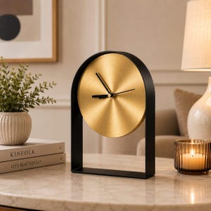 Puede incluir: Un reloj de mesa con una esfera circular de tono dorado cepillado y manecillas negras, encerrado en un marco de arco de metal negro mate. El reloj está diseñado para mantenerse en posición vertical sobre una superficie plana y tiene una silueta geométrica.