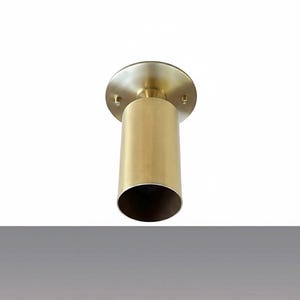 May include: A brass-colored, cylindrical ceiling light fixture. The fixture has a circular base with mounting holes and a downward-facing, open-ended cylinder. The light is designed to be mounted on a ceiling.
