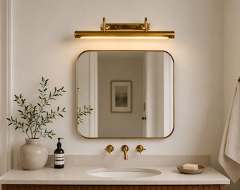 May include: A rectangular wall mirror with rounded corners and a thin gold frame, mounted above a bathroom vanity. A horizontal gold light fixture is positioned directly above the mirror, casting a warm glow downwards.