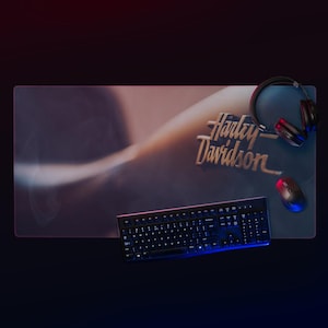 May include: A Harley Davidson-branded desk mat with a keyboard, mouse, and headphones. The mat has a gradient design with the Harley Davidson logo. The keyboard and mouse are black, and the headphones are black and red.