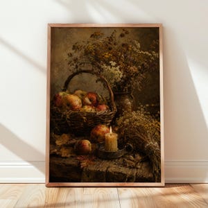 May include: A framed wall art print depicting a still life scene with a woven basket filled with red and yellow apples, a lit pillar candle in a metal holder, and dried wildflowers arranged in a vase, all set on a rustic wooden surface.