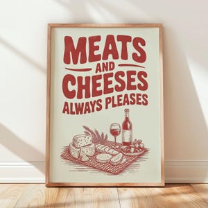 May include: A framed print with the words "MEATS AND CHEESES ALWAYS PLEASES" in red. The image below depicts cheese, bread, wine, and grapes. The print is in a wooden frame and leans against a white wall.