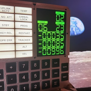 Puede incluir: Una réplica de la interfaz DSKY del ordenador de guiado Apollo con una rejilla de botones de estado blancos iluminados etiquetados como UPLINK ACTY, NO ATT, STBY, KEY REL, OPR ERR, TEMP, GIMBAL LOCK, PROG, RESTART, TRACKER, ALT y VEL, junto a una pantalla digital verde.