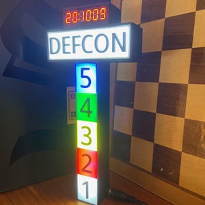 Może przedstawiać: Pionowy wskaźnik statusu DEFCON z cyfrowym zegarem na górze pokazującym 20:00:09. Poniżej tekstu znajduje się pięć ułożonych w stos prostokątnych paneli podświetlonych na niebiesko, zielono, żółto, pomarańczowo i czerwono, oznaczonych cyframi od 5 do 1.