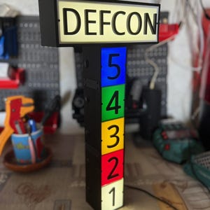 Può includere: Un'insegna rettangolare DEFCON con cornice nera e pannello superiore illuminato. Sotto il testo, una colonna verticale presenta cinque pannelli quadrati impilati e illuminati in blu, verde, giallo, rosso e bianco, con i numeri 5, 4, 3, 2 e 1.