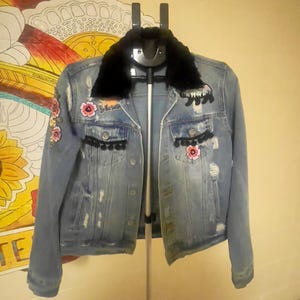Può includere: Giacca di jeans blu chiaro con colletto in ecopelliccia nera. La giacca presenta ricami floreali, finiture con pompon neri e dettagli strappati. Toppe ricamate sono visibili sulle spalle e sulle tasche. La giacca è appesa a una gruccia nera.