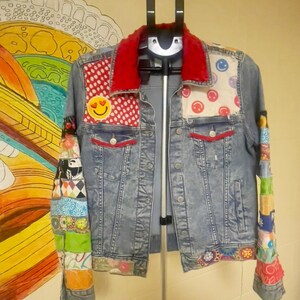 May include: A denim jacket with a red corduroy collar and patches of smiley faces and polka dots. The sleeves are adorned with colorful fabric patches. The jacket has red pocket trim and is displayed on a clothing rack.