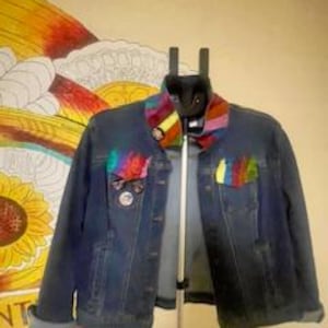 May include: A dark wash denim jacket with rainbow accents on the pockets and collar. The jacket is hanging on a black hanger against a wall with a colorful sunflower mural. The jacket has a button-up front and cuffed sleeves.