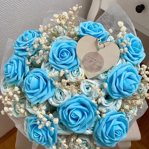 Puede incluir: Un ramo de rosas artificiales de color azul brillante intercaladas con flores blancas de paniculata. Una etiqueta de madera en forma de corazón con el texto "HANDMADE with love" está sujeta al centro del arreglo, envuelto en papel translúcido.