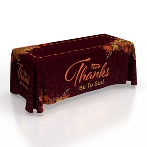 May include: A burgundy tablecloth with a fall leaf pattern and the words "Thanks Be To God" in orange script. The edges are decorated with orange and yellow leaves. The tablecloth is draped over a rectangular table.
