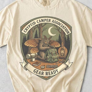 Może przedstawiać: Kremowy t-shirt z grafiką przedstawiającą stworzenie przypominające ćmę w lesie z ekwipunkiem kempingowym. Napis "CRYPTID CAMPER ASSOCIATION" oraz "GEAR READY" otacza półksiężyc i latarnię.