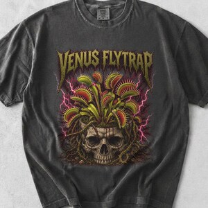 May include: A charcoal gray short-sleeve t-shirt featuring a graphic print of a human skull with green and red Venus flytrap plants growing out of the top, surrounded by pink lightning bolts and the text VENUS FLYTRAP in yellow lettering.