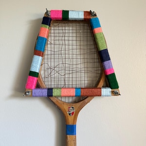 May include: A vintage tennis racket with a wooden frame and a wire mesh net. The frame is wrapped with colorful yarn in shades of pink, green, blue, orange, and purple. The handle is wooden with a blue grip and a shield emblem.