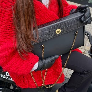 Könnte beinhalten: Eine schwarze Handtasche mit einem goldfarbenen Kettenriemen und einer strukturierten Klappe mit Krokodilmuster. Die Tasche wird von einer Person gehalten, die schwarze Handschuhe und einen leuchtend roten, grob gestrickten Pullover trägt.