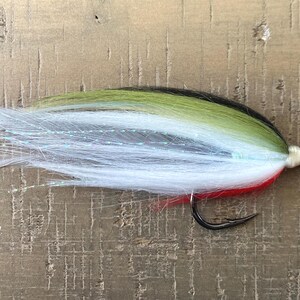 May include: A colorful fishing fly with a black hook. The fly features a combination of white, green, black, and red materials, designed to mimic bait. The fly is designed for attracting fish.