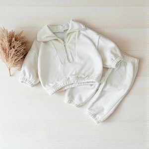 May include: A cream-colored, two-piece outfit for children. The set includes a long-sleeved sweatshirt with a zip-up collar and matching sweatpants. The outfit is made of a soft, textured material and is designed for comfort.