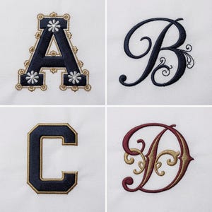 May include: Four embroidered letters on a white fabric background. The letter A is navy with gold trim and white flower accents. The letter B is navy cursive. The letter C is navy with a gold border. The letter D is burgundy with gold filigree.