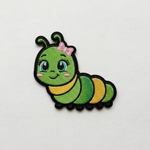 May include: Embroidered patch of a cartoon caterpillar with a smiling face, large blue eyes, and a pink bow. The caterpillar is mainly green with yellow and black details, suitable for decorating clothes or accessories.