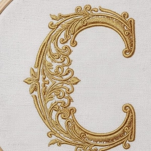 May include: Embroidered gold letter "C" with ornate, swirling floral details on a white fabric background. The letter is framed in a wooden embroidery hoop. The design features a classic, elegant style.