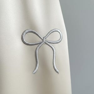 May include: Close-up of a cream-colored fabric with a silver embroidered bow. The bow is centered and detailed, with the fabric slightly textured. The background is a solid, neutral color.