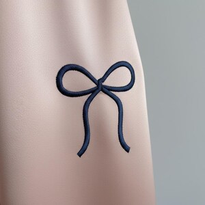 Puede incluir: Primer plano de una tela rosa pálido con un lazo bordado en azul marino. El lazo está centrado y tiene un diseño clásico anudado. La tela tiene una textura suave.