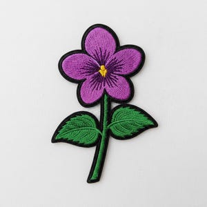 Puede incluir: Parche bordado de una flor morada con centro amarillo y contorno negro. La flor tiene cinco pétalos y está unida a un tallo verde con dos hojas verdes. El parche está diseñado para planchar o coser.