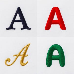 May include: Four embroidered letter A designs on a white fabric background. The top left is a navy blue serif font, the top right is a red sans-serif font, the bottom left is a gold cursive script, and the bottom right is a green rounded font.