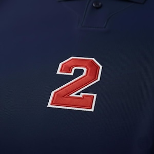 May include: Close-up of a navy blue polo shirt with a red number 2 patch. The number is outlined in white and has a textured, embroidered appearance. The shirt has a ribbed texture.