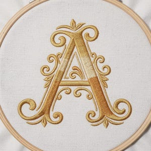 Peut inclure: Broderie de la lettre "A" en fil d'or sur tissu blanc, encadrée dans un cercle à broder en bois. Le motif présente des volutes et des ornements élaborés, créant un monogramme élégant.