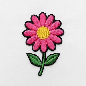 Puede incluir: Parche bordado de una margarita rosa con centro amarillo y contorno negro. La flor tiene hojas verdes y un tallo verde. El parche es probablemente para ropa o accesorios.