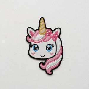 May include: Embroidered unicorn patch with a white face, pink and white hair, and a gold horn. The unicorn has blue eyes, a pink bow, and a black outline. This patch is suitable for clothing, bags, or other fabric items.