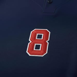 May include: Close-up of a navy blue polo shirt with a red and white embroidered number 8 patch. The number has a white border and is stitched with a textured pattern. The shirt has a buttoned placket.
