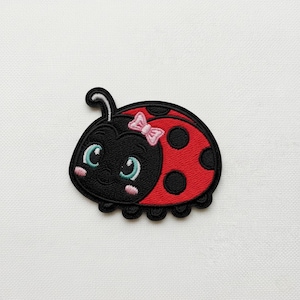 Op de afbeelding: Geborduurde lieveheersbeestjespatch met een zwarte kop, rood lichaam en zwarte stippen. Het lieveheersbeestje heeft een roze strik, blauwe ogen en een lachend gezicht. Voor kleding of accessoires.