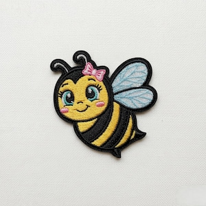 Könnte beinhalten: Gestickter Aufnäher mit einer Cartoon-Biene mit rosa Schleife, blauen Augen und gestreiftem Körper. Die Biene hat schwarz-gelbe Streifen, blaue Flügel und ein lächelndes Gesicht. Der Aufnäher befindet sich auf weißem Hintergrund.