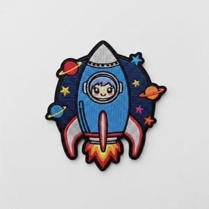 Puede incluir: Parche bordado con un cohete azul con un astronauta sonriente en la ventana. El cohete tiene detalles rojos y plateados, con llamas en la base. Planetas y estrellas rodean el cohete sobre un fondo azul oscuro.