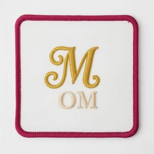 Puede incluir: Parche cuadrado blanco con borde burdeos. El parche presenta la letra "M" dorada bordada sobre las letras "OM" en color beige claro. El diseño está centrado en el fondo blanco.
