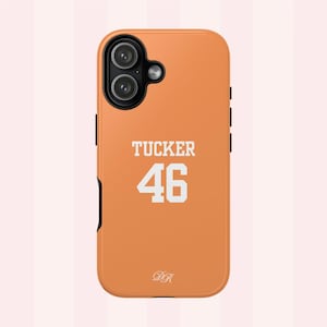 Può includere: Una cover per smartphone arancione con un ritaglio nero per il modulo fotocamera. Il retro della cover mostra il testo bianco TUCKER sopra il numero 46 in un carattere sportivo in grassetto, con un piccolo logo DR in corsivo bianco vicino al bordo.