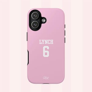 Può includere: Una custodia per smartphone rosa chiaro con un modulo fotocamera nero a due lenti. Il retro della custodia mostra il testo bianco LYNCH sopra il numero 6 in carattere stampatello, con le piccole iniziali bianche in corsivo DR in basso.