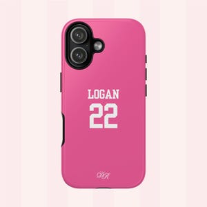 May include: A bright pink phone case featuring a black camera housing with two lenses and a flash. The back displays the white text LOGAN above the number 22 in a block font, with a small white cursive DR monogram near the bottom edge.
